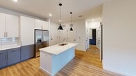 This thoughtfully designed one-bedroom, one-bathroom unit offers a functional and modern living experience across a single floor. The open-concept kitchen serves as the centerpiece, featuring a white quartz-topped island with an undermount sink, dual pendant lights in matte black, stainless steel appliances including a French door refrigerator with ice maker, a range, microwave, and dishwasher. White upper cabinetry pairs with gray lower cabinets, complemented by a classic subway tile backsplash. Warm-toned hardwood-style flooring flows throughout the main living and kitchen areas, adding a sense of continuity and warmth.

The bedroom is carpeted for comfort and includes a ceiling fan, a window with a roller shade that allows natural light to filter in, and a clean, neutral color palette. A secondary carpeted room, visible in the layout, provides additional flexible space.

The bathroom is well-appointed with a long gray vanity offering ample storage, a white quartz countertop, a large mirror, and wood-style flooring. A walk-in closet is accessible directly from the bathroom, enhancing storage capacity. A barn-style door adds a stylish architectural detail. The unit's crisp white walls and recessed lighting throughout create a bright, clean, and modern ambiance.
