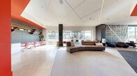 Located at 1912 S Jacaranda St in Anaheim, CA, this 1,190 square foot single-floor business center delivers a contemporary, open-plan environment designed for productivity and collaboration. The space is defined by a striking modern aesthetic, combining polished concrete flooring, patterned carpet tiles, and bold orange accent walls and ceilings that energize the interior. Large floor-to-ceiling windows span multiple walls, flooding the space with natural light and offering views of the surrounding greenery.

The main lounge area features a curved modular sofa in neutral gray, complemented by individual lounge chairs with red accents, creating versatile seating arrangements suited for both informal meetings and relaxed work sessions. A large wall-mounted flat-screen TV sits above a sleek, floating wood-and-white credenza with open shelving, serving as a media and resource hub.

A standout feature is the oversized street map mural adorning a full accent wall, adding an artistic, location-specific design element. The kitchenette area is equipped with a stainless steel refrigerator, microwave, and ample open shelving with gray cabinetry. Linear recessed lighting and LED strip fixtures on the ceiling provide consistent, even illumination throughout. Glass-partitioned sections offer semi-private work zones, rounding out this well-appointed, multifunctional business center.