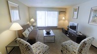 This single-floor residence features a welcoming living room as its centerpiece. The space is finished with wall-to-wall carpeting in a neutral tone, contributing to a warm and comfortable atmosphere. A large sliding glass door fitted with vertical blinds serves as the primary source of natural light, brightening the room and creating an open feel.

The living room is thoughtfully arranged with a dark upholstered sectional sofa accented by patterned throw pillows, complemented by two matching accent chairs with a circular geometric print. A sleek, rectangular coffee table in dark wood anchors the seating area and holds a decorative bowl as a focal point. Opposite the seating arrangement, a low-profile media console supports a flat-screen television, completing the entertainment setup.

Ambient lighting is provided by two floor lamps positioned on either side of the room, adding warmth during evening hours. A wall-mounted air conditioning unit ensures year-round climate control. The neutral-toned walls and textured ceiling lend the space a clean, understated aesthetic suited to a range of décor styles.