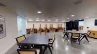 Located in the heart of Los Angeles at 1120 W 6th St, this thoughtfully designed single-floor workspace offers a versatile range of environments suited for focused work, collaboration, and relaxation. The main open area features polished concrete flooring and recessed lighting throughout, creating a clean and contemporary aesthetic. A row of built-in booth workstations lines one wall, each constructed from warm natural wood with cognac leather upholstered seating, providing semi-private work settings. Complementing these are multiple dark-wood tables paired with rattan-accented chairs, offering additional flexible seating arrangements. A glass-partitioned conference room offers a more enclosed meeting environment, fitted with a round table, cane chairs, and built-in wood shelving. Adjacent to the open workspace is a lounge area finished with light hardwood flooring, featuring a curved cognac leather sofa, plush white boucle chairs, a large wall-mounted display, and a decorative olive tree that adds warmth. A sleek kitchenette with warm wood cabinetry, marble countertops, a matte black faucet, and stainless steel appliances serves the space. The overall design blends natural materials with modern finishes, creating a professional yet inviting atmosphere.