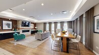 Located at 1551 E Central Rd in Arlington Heights, IL, this single-floor community clubhouse offers a thoughtfully designed amenity space that blends comfort, function, and modern style. The open-plan kitchen and living room serves as the heart of the facility, featuring warm hardwood flooring, a tray ceiling with recessed and cove lighting, and an abundance of natural light from large windows dressed with elegant draping curtains. The living area centers around a striking stone fireplace flanked by a wood-accented accent wall, a wall-mounted flat-screen TV, and vibrant teal accent chairs that add a pop of color to the neutral palette. A generous natural wood dining table with contemporary seating provides ample space for gatherings. The kitchen is outfitted with rich dark cabinetry, light quartz countertops, a glass tile backsplash, stainless steel appliances, and a breakfast bar with bar seating. Adjacent to the main lounge, the exercise room is bright and airy, boasting soaring ceilings, large floor-to-ceiling windows overlooking an outdoor pool and green surroundings, and a comprehensive selection of cardio and strength-training equipment, including treadmills, ellipticals, stationary bikes, and a multi-station weight machine.