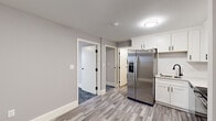 Located at 101 Cedar St in Providence, Rhode Island, this single-floor unit offers a thoughtfully updated interior with a clean, contemporary aesthetic throughout. The kitchen is a standout feature, showcasing white shaker-style cabinetry with matte black hardware, light-toned countertops, and a full suite of stainless steel appliances including a side-by-side refrigerator with ice and water dispenser, electric range, over-the-range microwave, and dishwasher. Light wood-look flooring runs through the kitchen area, complementing the neutral gray walls. Multiple rooms feature plush patterned carpet in a blue-gray tone, providing a comfortable and modern feel. Each carpeted room benefits from natural light through windows that frame views of the surrounding Providence cityscape, and baseboard heating ensures year-round comfort. One room also includes a wall-mounted air conditioning unit. The full bathroom is finished in a crisp, all-white palette with a single-sink vanity, white cabinetry, a tub-shower combination with white tile surround, and coordinating matte black fixtures and accessories. White trim and doors with black hardware tie the design together cohesively throughout the space, creating a polished and move-in-ready environment.