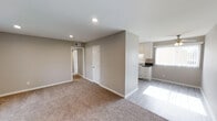 This single-floor space offers a practical, open-concept layout that seamlessly connects the living area to the kitchen. The living space is finished with wall-to-wall carpeting in a warm neutral tone, complemented by soft taupe-painted walls and clean white baseboards that create a cohesive, understated aesthetic throughout.

Recessed lighting in the ceiling provides even illumination across the main living area, while a visible HVAC vent ensures climate control. Two interior doors along the back wall lead to additional rooms, suggesting functional separation of private spaces from the main living area.

The kitchen, partially separated by a dividing wall, transitions to smooth hardwood-style flooring and features white cabinetry, a white range, and dark countertops. A window in the kitchen area allows natural light to filter into the space, brightening the area considerably.

Overall, the space maintains a clean, minimalist character with a neutral color palette, making it highly adaptable to various furnishing styles. The straightforward, efficient layout prioritizes functionality while maintaining a comfortable and welcoming atmosphere.
