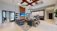 Located at 267 Nance Rd in Huntsville, Alabama, this single-floor community clubhouse offers a generously sized, thoughtfully designed gathering space that blends modern sophistication with warm, inviting elements. The main lounge serves as the centerpiece of the facility, showcasing soaring ceilings adorned with rich, dark wood beam detailing and dramatic circular pendant light fixtures. A floor-to-ceiling walnut wood accent wall anchors the room, featuring a large wall-mounted flat-screen TV flanked by open shelving. Plush seating arrangements, anchored by a patterned area rug over light-toned tile flooring, create an inviting atmosphere for socializing and relaxation. An adjacent semi-private lounge nook provides intimate booth-style seating beneath pendant lighting, ideal for small gatherings or quiet conversation. The entertainment wing transitions into a billiards area and media lounge, where bold teal accent walls, a geometric white wall feature, exposed dark beam ceilings, and a full-size pool table create a dynamic recreational atmosphere. A well-appointed kitchen island with white quartz countertops and leather bar seating anchors the catering area, which benefits from expansive windows offering outdoor views of the surrounding community. Natural light flows generously throughout, enhancing the cohesive, upscale aesthetic of this versatile community hub.