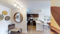This single-floor residence showcases a thoughtfully designed open-concept layout that connects the entryway, kitchen, and living areas into a cohesive, flowing space. Light hardwood-style flooring runs throughout, adding warmth and visual continuity across the unit.

The kitchen is a standout feature, equipped with rich dark wood cabinetry, stainless steel appliances including a range, microwave, and refrigerator, and a clean white subway tile backsplash. The light-toned countertops provide a striking contrast to the darker cabinetry. A peninsula-style breakfast bar with barstool seating extends into the living area, making it ideal for casual dining or entertaining.

The entryway area is tastefully appointed, featuring track lighting overhead and a decorative round wood-framed mirror that adds character to the space. Recessed lighting throughout ensures the interior remains bright and well-illuminated. White walls keep the overall aesthetic clean and contemporary, allowing the natural wood tones and stainless finishes to stand out. The open layout enhances the sense of space, making the unit feel airy and inviting.