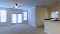 This single-floor space offers a clean, functional layout with a welcoming ambiance throughout. The living area is finished with plush wall-to-wall carpeting and features elegant crown molding that frames the ceiling, adding a refined architectural touch. A ceiling fan with an integrated light fixture is centrally positioned, providing both comfort and illumination. Natural light fills the room generously through a set of French doors fitted with blinds, which open to an outdoor area, and an additional window alongside them further brightens the space. The neutral, warm-toned walls create a versatile backdrop that complements the overall design. A partial wall with a raised countertop — finished in dark granite or stone — separates the living area from the adjacent kitchen, offering a convenient breakfast bar or pass-through. The kitchen area is partially visible, hinting at additional counter space beyond. The open-concept flow between the living and kitchen areas makes the space feel connected and spacious. White baseboards and trim throughout provide crisp contrast against the soft carpet and beige walls, contributing to the space's tidy, move-in-ready presentation.