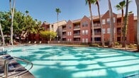 This two-story brick residential complex presents a classic architectural style defined by its warm red brick exterior, gabled rooflines, and private balconies that overlook the well-maintained grounds. The buildings are arranged to create an inviting courtyard atmosphere, with the centerpiece being a generously sized outdoor swimming pool featuring a connected spa. The pool area is framed by a paved deck outfitted with lounge chairs, metal fencing, and lush greenery, making it an ideal space for relaxation and recreation.

Towering palm trees and mature shade trees are distributed throughout the property, lending a resort-like ambiance and providing natural shade across the outdoor spaces. A paved walkway winds through the grounds, lined with manicured landscaping, garden lighting, and benches, encouraging leisurely strolls through the tree-canopied pathways. The interplay of dappled light through the palms and oaks adds a dynamic visual quality to the outdoor environment throughout the day.

The multi-building layout creates distinct zones for community amenities while maintaining a cohesive aesthetic. The brick facade, pitched rooflines, and covered balconies contribute to a traditional yet comfortable character, while the abundant greenery and outdoor amenities enhance the overall livability and appeal of the complex.