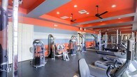 This fitness center makes a strong visual impression with its bold interior design, characterized by striking red and gray horizontal striped walls that create an energetic atmosphere suited for exercise. The ceiling continues the color scheme with vibrant red panels accented by gray beams, recessed lighting, and dual ceiling fans that help maintain airflow and comfort throughout the space. The room is generously sized, accommodating a comprehensive selection of strength training equipment, including multiple cable machines, weight stations, and resistance equipment arranged to allow ease of movement between stations. A floor-to-ceiling mirror spans one wall, enhancing the sense of space while allowing users to monitor their form during workouts. The dark rubberized flooring provides both durability and safety underfoot. Cardio equipment is also visible along the perimeter of the room, offering a well-rounded fitness experience. The combination of functional layout, ample equipment variety, and energetic design elements makes this fitness center a practical and motivating environment for users of all fitness levels.