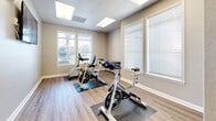 Located at 2901 Gandy Blvd N in Pinellas Park, Florida, this dedicated fitness room offers a focused and well-appointed exercise space designed for cycling workouts. The room is finished with durable wood-look luxury vinyl plank flooring throughout, providing a clean, low-maintenance surface ideal for active use. Multiple spinning bikes and a recumbent stationary bike are positioned on protective floor mats, offering a variety of cardiovascular workout options to suit different fitness levels. A wall-mounted flat-screen television is paired with the word "MOTIVATION" displayed prominently on the accent wall, creating an energizing atmosphere for guided workouts and on-screen cycling classes. The space is illuminated by flush-mounted panel lights set into a drop ceiling grid, supplemented by generous natural light streaming through several windows fitted with white horizontal blinds. The neutral-toned walls and white trim contribute to a clean, welcoming aesthetic. A small utility desk provides a functional surface for personal items. The single-story layout is efficiently organized, with a glazed entry door allowing visibility into and out of the space. Overall, this fitness room delivers a practical and motivating environment well-suited for community or facility use.