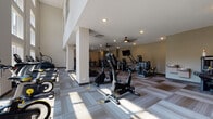 This two-story fitness center offers a well-appointed exercise environment designed to accommodate a wide range of workout routines. The space is finished with neutral-toned carpet tile flooring in a geometric pattern throughout, contributing to a clean and functional aesthetic. Recessed lighting and multiple ceiling fans are distributed across both levels, ensuring consistent air circulation and illumination throughout the facility.

The main floor features an extensive selection of cardio equipment, including treadmills, stationary bikes, ellipticals, and stair climbers, many from the True Fitness brand. Large windows and glass doors on this level allow natural light to fill the space and provide views of the surrounding outdoor area. A dedicated strength-training section includes multi-station cable machines, selectorized weight machines, a functional trainer, and a punching bag, offering options for both resistance and functional training.

A staircase connects the main floor to the upper level, which provides additional strength and conditioning equipment. Wall-mounted televisions are positioned throughout for entertainment during workouts. The entrance area features glass doors, a sanitization station, and informational signage, establishing an organized and welcoming entry point to the facility.