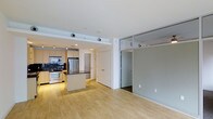 This single-floor Dallas residence offers a thoughtfully designed open-concept layout that seamlessly connects the living and kitchen areas. The contemporary kitchen is outfitted with stainless steel appliances, including a refrigerator, microwave, and stove, complemented by light wood cabinetry, a dark granite-topped island, and a bold tile backsplash that adds a striking visual contrast. Light hardwood flooring runs throughout the main living area, enhancing the sense of openness and warmth. A defining architectural feature is the frosted glass partition walls framed in sleek metal, which elegantly divide spaces while allowing light to flow freely and maintaining a bright, airy atmosphere. Recessed lighting in the white ceiling provides clean, even illumination throughout. The layout includes additional rooms accessible through white panel doors, offering flexible use of space. The overall ambiance is modern and minimalist, with clean lines, neutral tones, and a cohesive design language that gives the space a polished, urban feel well-suited to the dynamic Dallas lifestyle.