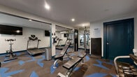 Located at 914 W Lake St in Fort Collins, Colorado, this dedicated fitness center offers a well-equipped exercise space across a single floor. The room is designed to maximize functionality, with a thoughtfully arranged layout that accommodates a diverse range of workout options.

The space features a full complement of cardio equipment, including treadmills and a stationary bike, each positioned beneath wall-mounted flat-screen TVs for an engaging workout experience. Strength training is well represented with a multi-station cable machine, a squat rack with a weight bench, adjustable dumbbells on a rack, and medicine balls of various sizes. An inversion table adds a recovery and flexibility dimension to the offerings.

Large mirror panels line portions of the walls, enhancing the sense of openness and allowing users to monitor their form. Recessed ceiling lighting provides bright, even illumination throughout. Frosted glass panels and interior windows contribute to a clean, modern aesthetic while maintaining a sense of visual connectivity within the space.

The floor is covered in a striking geometric-patterned carpet in shades of blue and brown, giving the gym a contemporary, energetic feel. Teal-accented structural columns punctuate the space, tying into the overall color scheme. A wall-mounted air conditioning unit ensures comfort year-round, and a stainless steel waste bin and hand sanitizer station reflect attention to cleanliness and convenience.