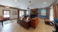 This single-floor space is thoughtfully designed to balance comfort and functionality, featuring two distinct yet connected areas: a living lounge and a full kitchen. The living area makes a strong visual impression with its bold, rust-orange sectional sofa accented by teal and patterned throw pillows, set atop a graphic area rug over light-toned ceramic tile flooring. A wood-trimmed fireplace with a built-in media niche serves as the focal point of the room, flanked by a slate-blue accent wall showcasing large-scale abstract artwork. Recessed lighting and a ceiling fan provide practical overhead illumination, while rust-colored drapery panels frame the windows, allowing natural light to filter in. A credenza near the center of the room provides additional surface space, and a dining table with chairs is positioned nearby for flexible use.

The kitchen is well-appointed with rich, dark cherry cabinetry, granite countertops, and a dark subway tile backsplash. Stainless steel appliances, including a refrigerator and built-in microwave, complement the space. A peninsula with seating for three bar-height chairs connects the kitchen to the adjoining area, while pendant lighting in warm amber tones adds a welcoming glow throughout.