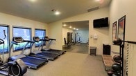 This single-floor fitness center offers a practical and well-organized space for a full range of exercise activities. The main area features multiple treadmills positioned along a windowed wall, allowing natural light to filter in and providing a pleasant view during cardio sessions. A wall-mounted flat-screen television adds an entertainment option for those using the cardio equipment. The space extends into a connected strength training zone, which houses a multi-function cable and weight machine, ideal for a variety of resistance exercises. The flooring transitions from carpet in the cardio area to a darker, rubberized surface in the strength training section, reflecting the functional design of each zone. Recessed lighting throughout ensures the space is well-lit, while a sanitizing station and waste bin are conveniently placed near the entrance, promoting cleanliness and hygiene. Additional amenities include a storage rack and a wooden bench for stretching or changing footwear. The light-colored walls and open layout contribute to an airy, welcoming atmosphere, making this fitness center a comprehensive and accessible facility for everyday use.