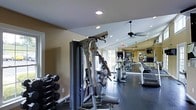 This fitness center offers a comprehensive workout environment designed to accommodate a wide range of exercise routines. The space features a dark, durable rubberized flooring that provides both safety and stability during workouts. A full-length mirrored wall runs along one side of the room, visually expanding the space and allowing users to monitor their form during exercise. Recessed lighting is embedded throughout the ceiling, ensuring the area is evenly and brightly lit, while a ceiling fan promotes air circulation during intense workouts. The equipment lineup includes a multi-function cable and weight machine, a row of treadmills, elliptical trainers, and a dedicated free weight station with a tiered dumbbell rack stocked with an assortment of weights. Natural light filters into the space through windows, adding to the open and energizing atmosphere. The warm-toned walls contribute to an inviting ambiance, while the thoughtful layout keeps equipment organized and accessible. An emergency exit sign is clearly visible, reflecting attention to safety standards. Overall, this fitness center is a well-appointed and functional space suited for both cardio and strength training.