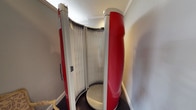 The tanning room is a purpose-built space designed to accommodate a full-size, stand-up tanning booth as its central feature. The booth itself is a cylindrical unit with a bold red exterior shell and a curved array of vertical tanning lamps lining its interior, accessed through a hinged door fitted with a chrome handle. The unit's circular footprint allows users to stand upright for an even, all-around tan, and a built-in control panel is mounted conveniently on the booth's exterior. The room's walls are finished in a neutral tone, and decorative crown molding along the ceiling adds a refined architectural detail that elevates the overall presentation of the space. The flooring is a dark carpet, providing a soft, comfortable surface underfoot. The room is compact and efficient, dedicated entirely to housing the tanning equipment without unnecessary clutter. The combination of the striking red booth against the neutral walls creates a visually distinct space. Overall, the tanning room offers a private, self-contained environment for indoor tanning, blending functional equipment with the polished interior finishes consistent with the broader property.