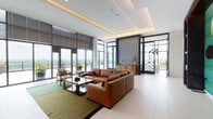 This thoughtfully designed single-floor amenity space blends contemporary aesthetics with functional comfort across several interconnected areas. The entrance corridor makes an immediate impression with large-format white tile flooring, a dramatic black-and-white chevron feature wall, and dark wood built-in shelving backed by a vivid teal textured panel. Patterned glass doors lead directly to an open-air terrace, framed by floor-to-ceiling windows that flood the interior with natural light and offer sweeping panoramic views of the surrounding landscape.

The main lounge area centers around a generous sectional sofa in warm cognac leather, anchored by a green area rug and a dark wood coffee table. Recessed tray ceilings with integrated ambient lighting add architectural depth overhead. Adjacent, a media lounge mirrors the warm palette, featuring another leather sectional, a wall-mounted television within a dark wood built-in unit, and a striking circular gold wall art installation against the teal accent wall.

A well-appointed kitchenette serves the space with white stone countertops, warm wood cabinetry, open metal-framed shelving, a stainless steel sink, and a built-in microwave—finished with a richly patterned geometric wood backsplash. Together, these elements create a polished, welcoming environment suited for both social gatherings and relaxed everyday use.