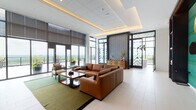 This thoughtfully designed single-floor amenity space blends contemporary aesthetics with functional comfort across several interconnected areas. The entrance corridor makes an immediate impression with large-format white tile flooring, a dramatic black-and-white chevron feature wall, and dark wood built-in shelving backed by a vivid teal textured panel. Patterned glass doors lead directly to an open-air terrace, framed by floor-to-ceiling windows that flood the interior with natural light and offer sweeping panoramic views of the surrounding landscape.

The main lounge area centers around a generous sectional sofa in warm cognac leather, anchored by a green area rug and a dark wood coffee table. Recessed tray ceilings with integrated ambient lighting add architectural depth overhead. Adjacent, a media lounge mirrors the warm palette, featuring another leather sectional, a wall-mounted television within a dark wood built-in unit, and a striking circular gold wall art installation against the teal accent wall.

A well-appointed kitchenette serves the space with white stone countertops, warm wood cabinetry, open metal-framed shelving, a stainless steel sink, and a built-in microwave—finished with a richly patterned geometric wood backsplash. Together, these elements create a polished, welcoming environment suited for both social gatherings and relaxed everyday use.