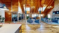 Located at 2751 Ruby River Dr in Lexington, Kentucky, this charming log cabin-style clubhouse delivers a warm, rustic aesthetic combined with modern amenities. The single-floor layout is defined by its most striking architectural feature: soaring vaulted ceilings with exposed timber beams and tongue-and-groove wood paneling that envelop the entire interior in rich, natural warmth. Wide-plank hardwood floors run throughout the main gathering area, complementing the handcrafted log wall construction.

At the heart of the lounge, a floor-to-ceiling stacked stone fireplace serves as a dramatic focal point, complete with a mounted television above. Multiple seating arrangements with plush blue upholstered chairs and sofas invite relaxed conversation, while a bar-height table and chairs provide an alternative gathering spot. Elegant chandelier lighting adds a refined touch overhead.

The fully equipped kitchen features dark cabinetry, white subway tile backsplash, stainless steel appliances, and a central island with seating — all beneath the same impressive vaulted ceiling flooded with natural light from clerestory windows. A dedicated package locker room with tile flooring and secure package lockers offers practical convenience. French doors open to a patio, seamlessly connecting the interior to the outdoors.