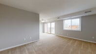 This single-floor residence offers a practical and well-organized layout across several distinct areas. The living room serves as the central gathering space, finished with neutral-toned wall-to-wall carpeting and soft gray walls that lend a clean, understated aesthetic. A ceiling fan provides both light and air circulation, while sliding glass doors open directly onto a private balcony with metal railings, allowing natural light to fill the room. A secondary window on an adjacent wall further brightens the space and provides additional outdoor views.

The kitchen is equipped with warm-toned wood cabinetry extending to the upper walls for generous storage, light-speckled countertops, and hardwood flooring. White appliances, including a refrigerator and dishwasher, complement the cabinetry and maintain a cohesive look.

The bedrooms are carpeted and finished in neutral gray tones, each benefiting from natural light through single windows fitted with blinds. One bedroom features a set of louvered bi-fold closet doors, while another includes a wall-length built-in closet system, offering substantial storage. A bathroom with tile flooring, a vanity with cabinet storage, and white fixtures rounds out the unit's functional offerings.