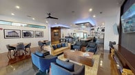 This single-floor communal space is thoughtfully designed to balance social gathering and focused work in a modern, urban aesthetic. The open-concept layout is anchored by polished concrete floors that reflect ambient lighting throughout, creating a seamless flow between distinct functional zones.

The main lounge area features a striking built-in electric fireplace framed by stacked firewood displays and a dark wood accent wall, with multiple large-screen TVs mounted above. Comfortable sectional seating and a substantial wood coffee table define the space, while recessed lighting and a ceiling fan maintain a relaxed atmosphere.

Adjacent to the lounge, a casual seating zone includes mid-century modern chairs and round dining tables, accented by a vertical wood slat partition and framed black-and-white architectural photography. Clerestory windows allow natural light to filter in along the upper walls.

The kitchenette is well-appointed with sage-green shaker cabinets, butcher block countertops, a built-in oven, a stainless refrigerator, and floor-to-ceiling open metal shelving functioning as a coffee bar. A billiards table occupies a dedicated area nearby, adding a recreational element.

A separate business center room features workstations, a wagon-wheel chandelier, a bold blue ceiling, and mid-century seating, offering a quiet retreat for productivity.