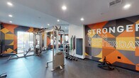 This modern fitness center is designed to inspire and motivate, combining a comprehensive equipment lineup with a bold, energetic aesthetic. The space is anchored by a striking large-scale mural in vivid shades of orange, yellow, and dark teal, emblazoned with the motivational phrase "Stronger Than Yesterday," which sets an uplifting tone throughout the entire floor.

The strength training area is thoroughly outfitted with Hoist cable machines, a functional trainer, a battle rope, resistance bands mounted to the wall, a set of kettlebells, and medicine balls on a dedicated rack. A padded workout mat provides space for floor-based exercises, while a wall-mounted flat-screen television and a built-in cabinetry counter area offer added convenience near the entry.

On the cardio side, a row of treadmills with integrated digital displays lines one wall, complemented by stationary cycling bikes. Full-length mirrors along the cardio area visually expand the space and allow users to monitor their form during workouts.

Large windows along the exterior walls flood the gym with natural light and provide views of the surrounding outdoor area, creating an open, airy atmosphere. The durable rubber flooring throughout provides both safety and functionality. Recessed lighting ensures the space is well-illuminated at all times, rounding out a fitness environment that is both practical and visually dynamic.