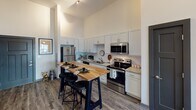 Located at 726 Fulton St in Chattanooga, Tennessee, this single-floor unit offers a thoughtfully designed open-concept layout that seamlessly connects the kitchen, dining, and living areas. The kitchen is appointed with white shaker cabinetry, granite countertops, subway tile backsplash, and a full suite of stainless steel appliances. A butcher block island with bar seating serves as both a functional workspace and a casual dining spot. Soaring ceilings throughout the main living area create an impressive sense of volume and openness, while durable wood-style flooring anchors the space. The living area is bathed in natural light from large windows that frame views of the surrounding streetscape, and a ceiling fan adds comfort overhead. The bedroom is a calm, well-proportioned retreat featuring plush carpeting, towering ceilings, and direct access to a full en-suite bathroom. The bathroom is finished with granite vanity countertops and wood-style flooring, and connects to a dedicated walk-in closet outfitted with built-in shelving and hanging storage. A wall-mounted flat-screen television and additional window complete the secondary bedroom space. Throughout, the unit balances contemporary finishes with a warm, livable ambiance.