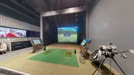 Located at 645 Division St in Nashville, TN, this single-floor indoor golf entertainment venue offers a modern, purpose-built environment designed for both serious golfers and casual visitors alike. The centerpiece of the space is a fully equipped golf simulator bay, featuring a large high-definition projection screen, a hitting mat with tee positions, interactive kiosk controls, and ball trays — providing an immersive simulation experience across a variety of virtual courses. Dark paneling frames the simulator screen wall, lending a polished, professional aesthetic to the bay.

Adjacent to the simulator is a comfortable lounge area furnished with stylish striped accent chairs, a wall-mounted flat-screen television, and a classic golf arcade cabinet tucked into an intimate alcove — perfect for those waiting their turn or simply relaxing between sessions. A beverage cooler with blue accent lighting adds a hospitality touch to this area.

Toward the rear of the space, exposed ductwork and industrial ceiling details contrast with the refined finishes below, giving the venue an urban, contemporary character. Bar-height tables and amber-cushioned stools line the back wall alongside a glass door entrance, creating a social gathering area. Lush potted plants add a refreshing natural element. A houndstooth-patterned accent wall ties the design together, balancing bold graphic detail with the space's overall cool, dark color palette.