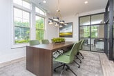 Located at 1401 W Paces Ferry Rd NW in Atlanta's prestigious Buckhead neighborhood, this single-floor professional space blends classic architectural details with contemporary design. The interior is defined by striking black steel-framed glass partition walls that divide the space into distinct functional zones while maintaining an open, transparent feel throughout.

Light hardwood flooring flows seamlessly across the entire floor, unifying the various areas. A conference or meeting room features a substantial dark wood table surrounded by green upholstered chairs, beneath a bold black-and-gold geometric pendant light. Tall, arched transom windows flood the room with natural light and offer views of the lush surrounding greenery.

Adjacent to the meeting room, an open collaborative lounge and kitchen area is anchored by a long marble-topped island with bar seating. This bright, airy zone is lined with large windows on multiple sides, enhancing the connection to the outdoors. Pendant lighting and fresh floral arrangements add warmth and sophistication.

Additional workstations and seating areas are visible through the glass partitions, offering flexible workspace configurations. Crown molding and recessed lighting throughout speak to the refined architectural character of the building, while the overall palette of soft whites and natural wood tones creates a professional yet welcoming atmosphere.