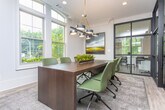 Located at 1401 W Paces Ferry Rd NW in Atlanta's prestigious Buckhead neighborhood, this single-floor professional space blends classic architectural details with contemporary design. The interior is defined by striking black steel-framed glass partition walls that divide the space into distinct functional zones while maintaining an open, transparent feel throughout.

Light hardwood flooring flows seamlessly across the entire floor, unifying the various areas. A conference or meeting room features a substantial dark wood table surrounded by green upholstered chairs, beneath a bold black-and-gold geometric pendant light. Tall, arched transom windows flood the room with natural light and offer views of the lush surrounding greenery.

Adjacent to the meeting room, an open collaborative lounge and kitchen area is anchored by a long marble-topped island with bar seating. This bright, airy zone is lined with large windows on multiple sides, enhancing the connection to the outdoors. Pendant lighting and fresh floral arrangements add warmth and sophistication.

Additional workstations and seating areas are visible through the glass partitions, offering flexible workspace configurations. Crown molding and recessed lighting throughout speak to the refined architectural character of the building, while the overall palette of soft whites and natural wood tones creates a professional yet welcoming atmosphere.