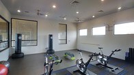 This single-floor fitness studio is thoughtfully designed to support a wide variety of workout routines in a clean, modern environment. The space features smooth, light-colored walls accented by bold black-framed windows that allow natural light to filter in, creating an energizing atmosphere. Recessed lighting and dual ceiling fans ensure the room remains well-lit and comfortably ventilated during intense exercise sessions. The flooring transitions between rubber fitness matting and carpet tile zones, providing appropriate surfaces for different types of activity. Equipment includes multiple stationary cycling bikes positioned for group spin classes, a freestanding punching bag for combat-style training, and step aerobic platforms for cardio and strength routines. Along one wall, a wall-mounted ballet barre offers support for stretching, balance training, and barre-style workouts. Pegboard panels line portions of the walls, offering practical storage potential for additional fitness accessories. The layout is open and uncluttered, maximizing usable floor space and allowing for flexible class arrangements or individual training. Overall, the studio presents a versatile, well-appointed environment suited for cycling, cardio, functional fitness, and low-impact training disciplines.