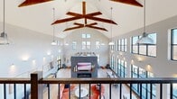 Located at 9 Monticello Drive in Brewster Village, NY, Westview is a striking two-story community amenity space designed with a blend of modern farmhouse aesthetics and contemporary sophistication. The main floor is expansive, anchored by a dramatic vaulted ceiling adorned with bold, exposed dark-wood beams that radiate outward in a sunburst pattern, flooding the space with architectural character. A double-height wall of black-framed floor-to-ceiling windows wraps the living area, inviting abundant natural light and offering sweeping views of the surrounding landscape and outdoor deck.

The main living area features a bold slate-clad fireplace with a mounted TV, surrounded by vibrant lounge seating and casual dining tables. A well-appointed kitchenette branded with the "Westview" name includes navy cabinetry, pendant lighting, and a bar-height communal counter — ideal for social gatherings. Additional seating nooks, a shuffleboard table, built-in teal bookshelves, and curated equestrian-themed artwork contribute to a warm, community-oriented atmosphere.

The upper floor offers a mezzanine-level overlook, providing a bird's-eye view of the soaring great room below. Hallways and entry areas feature textured plaid wallcovering, a package concierge station, and light wood flooring throughout, creating a cohesive and polished experience across both levels.