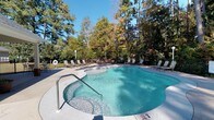 Located at 8132 Gayfer Road Ext in Fairhope, Alabama, this single-level outdoor amenity space centers around a generously sized freeform swimming pool with clear, blue-green water. The pool's curved, organic shape is complemented by a wide concrete deck that provides plenty of room for movement and relaxation. Numerous lounge chairs and seating options are arranged around the perimeter, offering comfortable spaces for sunbathing and socializing. Safety features, including a life ring station and clearly marked depth indicators, are thoughtfully integrated throughout the area. The space is enclosed by a perimeter fence and is framed by a striking backdrop of tall, mature trees — a mix of pines and hardwoods displaying rich seasonal color. The lush, wooded surroundings create a private, retreat-like atmosphere, effectively buffering the pool area from the outside environment. Natural light floods the space, enhancing the inviting quality of the water and the overall ambiance. The combination of the freeform pool design, generous deck space, and the enveloping natural canopy makes this outdoor area a standout feature, offering a tranquil and well-appointed setting for recreation and leisure.