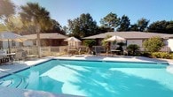 Located at 6413 Ashborough Ct in Milton, Florida, this single-story property offers a welcoming outdoor environment defined by its centerpiece swimming pool and thoughtfully arranged leisure spaces. The pool area showcases clear, blue water surrounded by a generously sized concrete deck, providing ample room for relaxation and recreation under the Florida sun. Multiple umbrella-shaded seating arrangements, including dining-height tables with chairs and reclining lounge chairs, are positioned throughout the deck, offering both shaded retreats and open sunbathing options. A classic white picket fence encloses the pool area, contributing a clean, tidy aesthetic while maintaining an open and airy feel. Tall palm trees and mature native trees frame the space, providing natural shade and a distinctly Floridian character. The surrounding landscaping includes tropical plantings that add texture and greenery to the environment. A flagpole displaying the American flag stands prominently nearby, adding a sense of place and community. The low-profile rooflines of the adjacent structures are visible in the background, reinforcing the single-story, accessible layout of the overall property. The combination of outdoor amenities, natural light, and verdant surroundings creates a relaxed, resort-like atmosphere well-suited to the warm Northwest Florida climate.