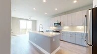 This well-appointed one-bedroom, two-bathroom residence offers a thoughtfully designed open-concept layout with modern finishes throughout. The kitchen serves as the centerpiece of the living space, showcasing a large central island with a white quartz countertop and undermount sink, sleek gray flat-front cabinetry, a subway tile backsplash, and a full suite of stainless steel appliances including a gas range, microwave, refrigerator, and a built-in wine cooler. Light wood-style flooring flows throughout the main living area, which is bathed in natural light and offers direct access to a private balcony through a large sliding glass door, with a ceiling fan adding comfort overhead. The bedroom features plush carpeting, a ceiling fan, and expansive windows that frame open views of the surrounding area, filling the room with abundant natural light. A walk-in closet with built-in shelving and hanging rods provides excellent storage. Both bathrooms are finished with large-format tile flooring, gray vanity cabinetry, white countertops, and frameless glass shower enclosures. A stacked washer and dryer unit is conveniently located near the primary bathroom, adding everyday practicality to this stylish and functional residence.