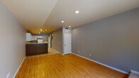 This two-floor space offers a well-organized layout with distinct living, kitchen, and sleeping areas. The main level features an open living area finished with warm hardwood flooring and neutral gray walls, illuminated by recessed ceiling lighting. Adjacent to the living space, the kitchen is equipped with white cabinetry, tile countertops, a full suite of white appliances including a refrigerator, electric range, built-in microwave, and dishwasher, all set atop light ceramic tile flooring. Track lighting adds additional brightness to the kitchen zone. A dedicated laundry area with a washer and dryer is conveniently located on this level. A bathroom with tile flooring, a vanity with storage, and a toilet is also accessible on the main floor. The upper level hosts carpeted bedrooms, each painted in warm taupe tones and featuring windows that allow natural light to filter in. The consistent use of neutral tones throughout both floors creates a cohesive and functional environment. Overall, the space balances practical amenities with a clean, understated aesthetic across its two levels.