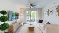 Located at 12171 Treeline Ave in Fort Myers, Florida, this single-floor, three-bedroom, three-bathroom home offers a clean, coastal-modern aesthetic defined by light tones, wood-look flooring, and an airy, well-organized layout. The open kitchen is a standout feature, boasting white shaker cabinetry extending to the ceiling, granite countertops, a tile backsplash, stainless steel appliances including a French door refrigerator, and a center island with bar seating — ideal for both cooking and casual dining. Track lighting overhead adds a contemporary touch. The living room is bright and welcoming, anchored by a ceiling fan, light-toned hardwood flooring, and sliding glass doors that open to an outdoor patio, framing lush greenery views. White walls and sheer drapes enhance the sense of openness. The bedrooms are comfortably scaled, each featuring plush carpeting, natural light from well-positioned windows, and a calm, neutral palette. The primary bedroom includes closet access supported by wire shelving systems. The bathrooms are finished with marble-look tile surrounds in the tub-shower combinations and granite vanity countertops, maintaining the home's cohesive, upscale design language throughout.