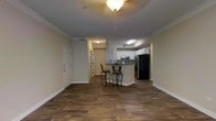 This single-story space showcases an open-concept layout that seamlessly connects the living area to the kitchen. Dark hardwood-style flooring runs throughout, providing a warm, cohesive foundation that contrasts cleanly with the neutral beige walls and bright white trim and baseboards. Crown molding lines the ceiling, adding a refined architectural detail throughout the main living space.

The kitchen is positioned toward the rear and features white cabinetry, dark countertops, and stainless steel appliances alongside a black refrigerator. A peninsula-style breakfast bar extends into the living area, accompanied by two decorative iron bar stools, creating a casual dining and entertaining space. The kitchen ceiling features a tray ceiling detail with a recessed light, distinguishing it from the main living area.

Near the entry, a light fixture with a frosted glass shade illuminates the space. The front door is flanked by a closet, offering convenient storage upon entry. A hallway leads further into the unit, suggesting additional rooms beyond the main open area. Overall, the space presents a practical yet polished interior with consistent design choices throughout.