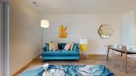 This single-floor residence offers a thoughtfully arranged layout across one bedroom and one bathroom. The living area serves as the heart of the space, featuring light wood-style flooring, neutral white walls, and a sliding glass door that opens to a private balcony, allowing natural light to fill the room. The open-concept kitchen is outfitted with dark cabinetry, stainless steel appliances including a refrigerator and microwave, pendant lighting, and a white peninsula countertop with bar seating for two — ideal for casual dining. A dedicated dining area sits adjacent to the kitchen, providing additional meal and entertaining options. The bedroom is generously proportioned, featuring the same warm wood-style flooring, crisp white walls, and ample space for a full dresser and seating area. A large framed artwork anchors the room above the bed, adding a vibrant focal point. The bathroom includes a full tub-shower combination with white tile surround, a vanity, and neutral tile flooring. Throughout the residence, clean lines, contemporary finishes, and a cohesive color palette create a modern and welcoming ambiance.