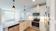This contemporary single-floor space showcases a thoughtfully designed open-concept layout that seamlessly connects the kitchen and living areas. The kitchen is a standout feature, outfitted with crisp white shaker-style cabinetry that extends to the ceiling, providing generous storage throughout. A full suite of stainless steel appliances includes an electric range, over-the-range microwave, refrigerator, and dishwasher. Light quartz countertops and a classic white subway tile backsplash contribute to the clean, modern aesthetic. A peninsula island with an undermount sink serves as a functional centerpiece, while a pair of industrial-style pendant lights add warmth and character overhead. Light wood-tone flooring runs throughout the main living areas, complementing the neutral color palette. The bedroom offers plush carpeting and a large window dressed with horizontal blinds, allowing for abundant natural light and views of the surrounding area. The bathroom is finished with white shaker vanity cabinetry, a gray countertop with undermount sink, a large mirror with open shelving, and a walk-in shower. Overall, the space presents a fresh, move-in-ready atmosphere with consistent modern finishes throughout.