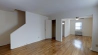 This two-floor space offers a clean, functional layout characterized by warm hardwood floors that run throughout the interior, reflecting natural light and adding a timeless quality to the overall aesthetic. The neutral white walls provide a versatile backdrop, creating a bright and open atmosphere across the living areas.

A standout feature is the built-in closet with rod storage, offering practical organization within the main living space. The open passage between rooms encourages a natural flow, with the adjoining area benefiting from a ceiling fan that aids in air circulation and comfort. Windows along the outer walls allow natural light to filter in, contributing to the airy feel of the space.

Trim details and baseboards are clean and consistent throughout, reinforcing the well-maintained character of the interior. A radiator is visible along one wall, indicating a classic heating system well-suited to year-round comfort. The staircase, partially visible, connects the two floors and hints at additional living space above.

Overall, this space presents a straightforward and functional design with durable finishes, ample natural light, and thoughtful storage solutions that make it both practical and inviting for everyday living.