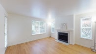 This well-maintained single-floor home offers a clean, modern aesthetic defined by crisp white walls, light hardwood flooring, and an abundance of natural light throughout. The spacious living room serves as the heart of the home, anchored by a classic tile-surround fireplace with a white-painted mantle. Large windows allow generous natural light to fill the room, while a glass-panel exterior door provides convenient outdoor access and further brightens the space. The galley-style kitchen is thoughtfully designed with white shaker cabinetry, dark countertops, and a full suite of stainless steel appliances, including a refrigerator, electric range, microwave, and dishwasher. A window above the sink brings in additional natural light. Adjacent to the kitchen, a charming bay window nook adds architectural interest and creates an inviting dining or sitting area. The layout also includes a bathroom with access to a carpeted bedroom area featuring a walk-in closet. The consistent hardwood flooring in the main living areas provides warmth and cohesion throughout the space, while the neutral color palette creates a versatile backdrop suited to a variety of interior styles.
