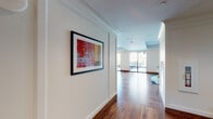 This expansive single-floor space is thoughtfully designed to offer a variety of functional zones, unified by warm-toned hardwood flooring and a consistently refined aesthetic throughout. A welcoming hallway introduces the space, accented by crown molding, crisp white walls, and a bold abstract artwork that sets an artistic tone.

The main gathering area is generously proportioned, featuring tray ceilings with ambient lighting and floor-to-ceiling windows that flood the interior with natural light and offer views of the surrounding landscape. A comfortable lounge zone presents plush seating arrangements, while a tufted green banquette booth with cafe-style tables provides a distinctive dining nook.

A dedicated recreation area centers on a full-sized billiards table beneath a brass pendant light fixture, complemented by large glass doors that open to an outdoor terrace. Adjacent to this, a formal dining area with upholstered seating offers additional gathering space.

The gourmet kitchen is a standout feature, showcasing rich walnut cabinetry, a substantial marble-topped island with leather barstools, and a striking sculptural brass chandelier. The overall ambiance is modern, polished, and versatile, designed to accommodate both social and leisure activities with equal elegance.