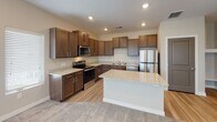 This single-story residence is designed with a practical, open-concept layout that connects the kitchen and living areas into one cohesive space. The kitchen serves as the centerpiece of the home, showcasing rich dark-brown cabinetry with brushed nickel hardware, light granite countertops, and a full suite of stainless steel appliances including a range, microwave, dishwasher, and refrigerator. A central kitchen island with matching granite provides additional workspace and a natural transition into the adjacent living area. Light wood-tone flooring runs throughout the kitchen, while plush carpet extends into the living and bedroom areas, adding warmth underfoot. Recessed lighting and a textured ceiling are consistent throughout the main areas. The living room benefits from multiple windows fitted with white blinds, allowing natural light to fill the space. Bedrooms are carpeted and feature ceiling fans and windows for ventilation and light. A dedicated laundry closet houses a full-size washer and dryer. The bathroom includes a full tub-shower combination with tile surround, standard fixtures, and tile flooring, maintaining a clean and functional aesthetic throughout.