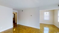 This single-floor space presents a straightforward, functional layout characterized by its warm, honey-toned parquet hardwood flooring, which adds a classic touch throughout the main living area. The floors are well-maintained and reflect light effectively, contributing to a bright overall atmosphere. The walls are finished in clean white, providing a neutral backdrop that enhances the sense of openness. A visible HVAC vent is integrated into the upper wall, indicating a central air system. White baseboards frame the perimeter of the room, offering a crisp architectural detail. A doorway on the left side of the main area leads to an adjoining hallway or secondary room, where an overhead light fixture is visible, suggesting continued living space beyond. To the right, a window allows natural light to filter into what appears to be an additional room or alcove, further brightening the interior. The layout feels open and connected, with a straightforward flow between spaces. The textured white ceiling adds subtle visual interest while maintaining a clean, modest finish. Overall, the space offers a well-lit, classic interior with durable flooring and a simple, functional design.
