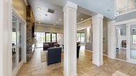 Located at 200 S Mallory St in Hampton, Virginia, this single-floor community clubhouse at Monroe Gates offers a thoughtfully designed gathering space that blends comfort with contemporary style. The entrance hallway greets visitors with floor-to-ceiling glass doors and sidelights, marble-look tile flooring, and soaring ceilings that fill the space with natural light. Elegant white wainscoting and soft gray walls establish a clean, refined aesthetic throughout. The spacious living room serves as the heart of the clubhouse, featuring a striking floor-to-ceiling stacked stone fireplace with a mounted TV, recessed lighting with crown molding, and large glass doors that open to outdoor views of the pool area. Plush leather seating, a patterned area rug, and decorative columns create a warm and inviting atmosphere. The game room stands out with its dramatic vaulted ceiling, a rustic chandelier, hardwood-style flooring, classic wainscoting, and a centerpiece billiards table framed by large windows. The kitchen is equally impressive, showcasing rich dark wood cabinetry extending to the ceiling, granite countertops, a large central island with bar seating, stainless steel appliances, and a trio of geometric pendant lights overhead — making it an ideal space for community gatherings and events.