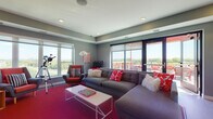 This single-floor space is thoughtfully designed as a contemporary lounge and gathering area, combining bold visual design with a functional, social layout. The interior is anchored by a generously sized living area featuring a large sectional sofa in charcoal gray, accented with vivid red and patterned throw pillows. A striking crimson carpet grounds the seating zone, complemented by a sleek white coffee table and a mix of mid-century-style armchairs in coordinating tones.

Large windows on multiple walls flood the space with natural light and frame sweeping elevated views of the surrounding landscape and greenery. A set of glass doors leads directly to a private outdoor terrace, enclosed by striped gray paneled walls and furnished with round bistro tables and bold red lattice chairs beneath string lights — ideal for outdoor gathering.

Back inside, one wall features a dramatic gradient orange-to-pink accent finish, adding an energetic focal point to a dedicated dining nook with a round table and upholstered chairs. A compact wet bar and beverage center are nearby. A telescope positioned near the corner windows adds a unique, leisure-oriented touch. Recessed ceiling lighting and a tray ceiling detail complete the polished, lively ambiance throughout.