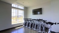 Located at 4115 Britton Parkway in Hilliard, Ohio, this fitness facility is designed to support an active and health-focused lifestyle. The space is single-story and thoughtfully laid out to maximize functionality for its users.

The standout feature of this facility is its dedicated spin studio, which is outfitted with a full row of stationary cycling bikes positioned for group class-style workouts. A large, wall-mounted flat-screen display faces the bikes, providing an ideal setup for instructor-led sessions or visual programming. The studio benefits from an expansive floor-to-ceiling window that floods the room with abundant natural light while offering open views of the surrounding outdoor landscape. The high placement of the windows adds to the sense of vertical space within the room, creating an airy and energizing atmosphere.

The interior finishes are clean and modern, featuring smooth white walls, polished concrete flooring, and a neutral color palette that contributes to a focused workout environment. The visible HVAC vents in the ceiling suggest a well-ventilated space, ensuring comfort during high-intensity exercise sessions.

Overall, this facility presents a well-equipped, bright, and professionally designed environment suitable for cycling classes and fitness training in the Hilliard community.