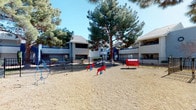 This outdoor communal space serves as a dedicated dog park and recreational area situated within a residential complex. The grounds are finished with a sandy, natural surface that provides a comfortable environment for pets to roam and play freely. The area is fully enclosed by a black wrought iron-style fence, ensuring a secure space for animals and their owners.

Several colorful pet agility and play structures are distributed throughout the space, including red horse-shaped spring riders, a red tunnel, and additional blue equipment, offering engaging activities for dogs of varying sizes. A mature pine tree stands prominently at the center of the area, providing generous natural shade across the grounds on sunny days. Additional seating, including blue benches, is available for residents to relax while supervising their pets.

Cornhole boards positioned near the perimeter suggest the area also serves as a casual recreational space for residents. The surrounding multi-story residential buildings feature a stucco exterior in neutral and blue tones, framing the space on multiple sides. The overall atmosphere is open, active, and community-oriented, designed to encourage outdoor enjoyment for both residents and their pets.