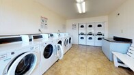 Located at 986 Armfield Circle in Norfolk, Virginia, this single-floor shared laundry facility is designed to serve the practical needs of its users with a clean, functional layout. The space is outfitted with a generous row of front-loading washing machines along one wall and a bank of stacked dryers along the opposite wall, providing ample capacity for multiple users simultaneously. Instructional signage is posted throughout to assist with machine operation. The flooring throughout is composed of neutral-toned tile, offering durability and ease of maintenance. White panel walls and overhead fluorescent lighting create a bright, well-lit environment. A white wooden bench is positioned along the wall, providing a comfortable waiting area for residents. A built-in counter surface is also available for folding or sorting laundry. Large glass doors and windows at one end of the room flood the space with natural light and offer a view of an enclosed outdoor courtyard, defined by brick walls and a railed upper walkway with mature trees visible beyond. The courtyard is accessible directly from the laundry room, adding a pleasant sense of openness to the otherwise utilitarian space. Overall, the facility is well-maintained and thoughtfully laid out to accommodate everyday laundry needs efficiently.